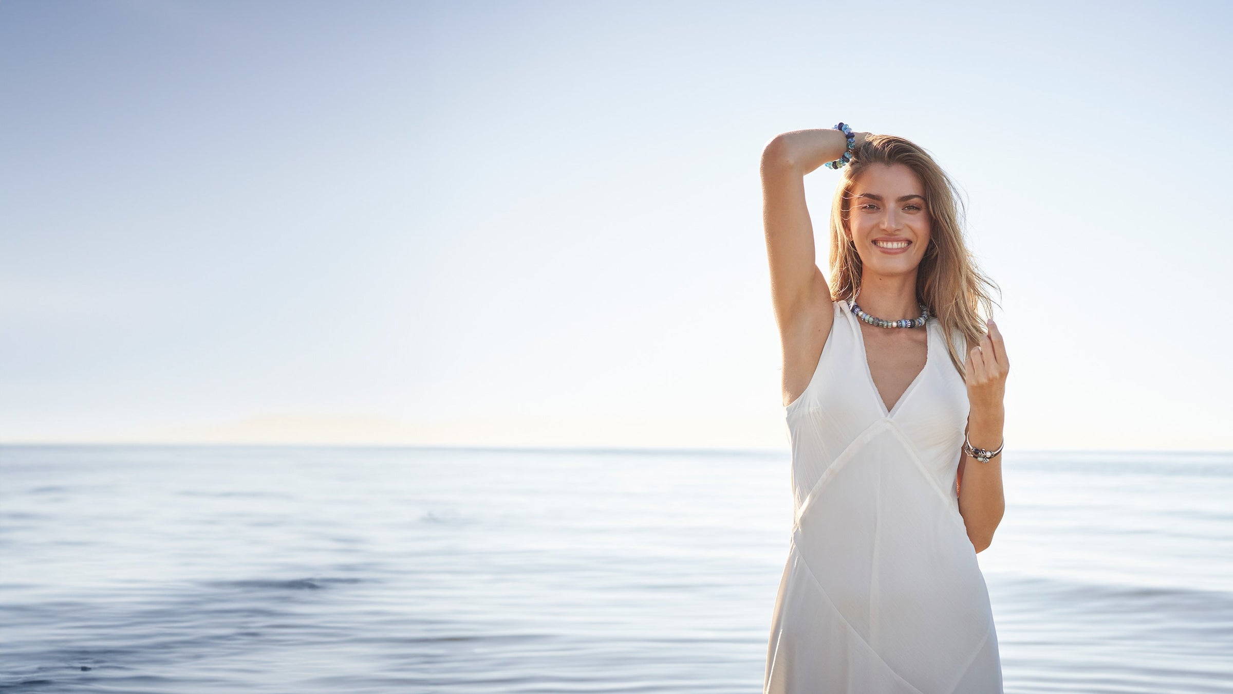 Modelwearing Trollbeads full glass bead necklace and bracelet, standing in the water at a beach smiling.